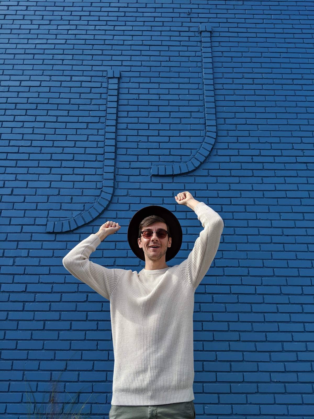 Jeff in a white sweater and hat, arms raised in front of a blue brick wall with a large JJ outlined in the brick